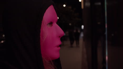 Woman-wearing-creepy-mask-and-hood-and-watching-a-flickering-tv-screen-at-night-in-urban-environment