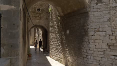 Dos-Personas,-Una-Mujer-Y-Un-Hombre-Caminando-Por-Los-Callejones-Del-Casco-Antiguo-De-Sibenik-Bajo-Un-Túnel-En-Un-Callejón-Estrecho-Protegido-Por-La-Sombra-En-Un-Caluroso-Y-Soleado-Día-De-Verano