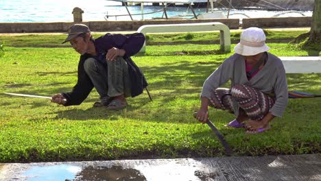 Two-gardeners-keep-the-grass-short-using-machetes-along-the-boulevard-of-Cagbianao,-Dinagat,-Philippines