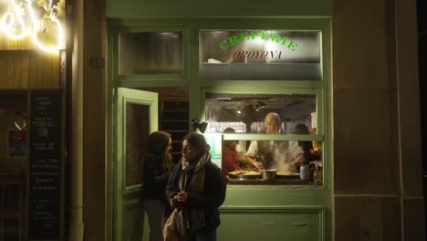 People-walking-in-and-out-a-Parisian-Creperie,-with-man-making-the-crepes-inside-in-Paris,-France