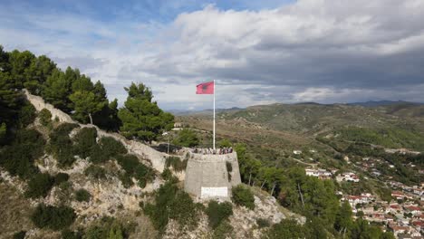 360-aerial-view-of-Albania-national-flag-waving-at-a-historical-building,-Barat-city-view-and-scenic-landscape-under-an-overcast-sky,-Europe