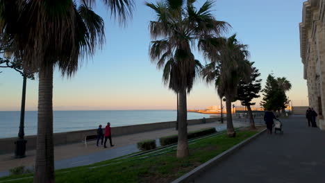 Paseo-Marítimo-Con-Palmeras-Al-Atardecer,-Gente-Caminando-Y-Un-Mar-En-Calma-Más-Allá