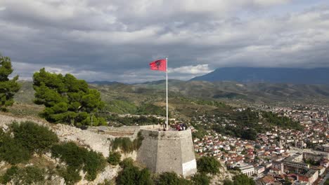 Toma-Cinematográfica-De-Un-Dron-De-La-Bandera-Nacional-De-Albania-Ondeando-En-Un-Edificio-Histórico-Con-Vistas-A-La-Ciudad-De-Barat-Y-Un-Paisaje-Escénico-Bajo-Un-Cielo-Nublado,-Europa