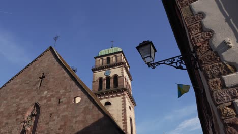 Iglesia-De-Sainte-Croix-En-El-Pueblo-Medieval-De-Kaysersberg-Con-Torre-Del-Reloj-En-Un-Día-Claro