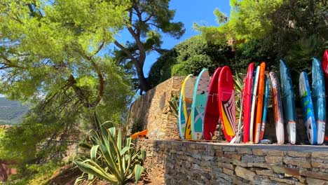 Colección-Colorida-De-Tablas-De-Charco-Junto-Al-Mar-En-Un-Lugar-Tropical-En-El-Sur-De-Francia-Con-árboles-Verdes-Y-Cielo-Azul,-Divertida-Actividad-Deportiva-De-Vacaciones-Soleadas,-Toma-De-4k