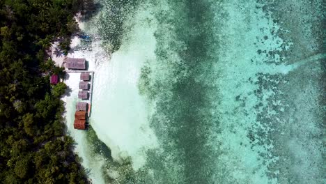 Aerial-view-of-remote-beach-huts-overlooking-stunning-crystal-clear-ocean-water-and-coral-reef-on-exotic,-remote-tropical-island-destination-in-Raja-Ampat,-West-Papua,-Indonesia