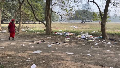 Sideview-of-a-sadhu-wearing-red-Indian-attire-and-walking-through-pathway-of-maidan-with-some-plastic-wastes-piled-up-near-it-in-Kolkata,-India