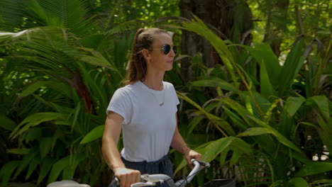 Una-Joven-Viajera-Con-Gafas-De-Sol-Caminando-Entre-Palmeras-Con-Su-Bicicleta-Hasta-La-Playa.