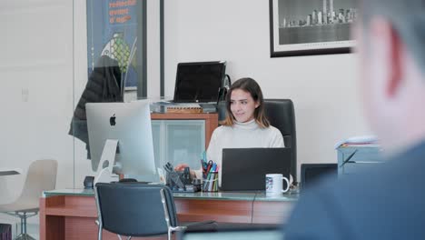 Young-female-teacher-sitting-behind-desk-on-her-laptop,-while-students-work-individually-on-assignment