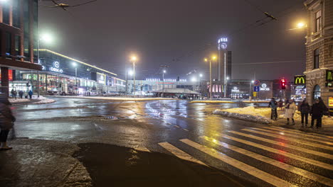 Toma-De-Timelapse-De-Peatones-Cruzando-La-Intersección-De-Calles-De-La-Ciudad-A-Lo-Largo-De-Caminos-Mojados-Causados-Por-El-Derretimiento-De-La-Nieve-Durante-La-Noche