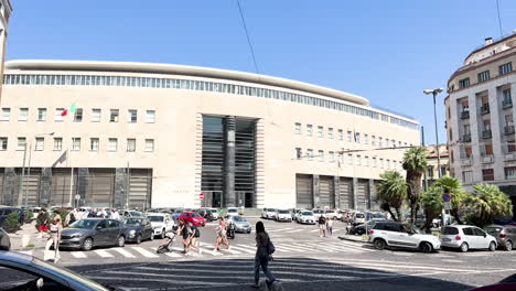 A-modern-building-in-Naples-with-a-curved-façade,-large-vertical-windows,-and-Italian-flags,-juxtaposed-against-a-busy-street-scene-with-pedestrians-and-vehicles