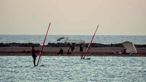 Windsurfer-Bereiten-Ihre-Ausrüstung-In-Der-Abenddämmerung-Auf-Einem-Ruhigen-Meer-Mit-Strandhintergrund-Vor