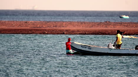 Two-people-in-bright-clothes-on-small-boats-by-the-shore,-with-clear-blue-water-and-a-distant-landmass