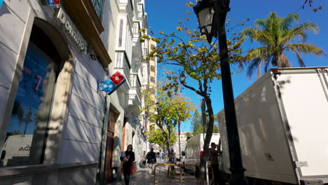 A-quaint-Cádiz-street-featuring-traditional-architecture-with-a-bright-advertisement-banner,-against-a-backdrop-of-blue-sky-and-treetops