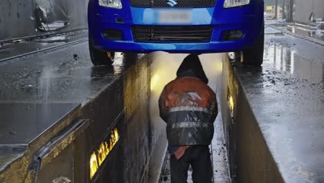 Mecánico-Con-Impermeable-Limpiando-Un-Coche-Desde-Abajo-Con-Una-Manguera-De-Agua-A-Presión-En-Un-Almacén-De-Limpieza-De-Coches