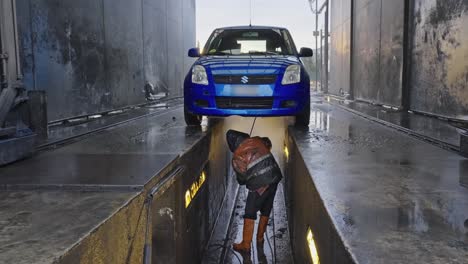 Persona-Limpiando-El-Tren-De-Aterrizaje-De-Un-Vehículo-Azul-Usando-Una-Manguera-De-Agua-A-Alta-Presión-Mientras-Está-Adornada-Con-Un-Impermeable-Protector-En-Una-Instalación-De-Limpieza-De-Automóviles.