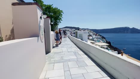 Pareja-Caminando-Mirando-Por-Encima-Del-Acantilado-|-Oia-Santorini-Grecia-Isla-Griega-En-El-Mar-Egeo,-Viajes-Turísticos-Vacaciones-Inmersivas-Caminatas-En-Movimiento-Junto-A-Multitudes-De-Compras-En-Acantilados-De-Mármol-Blanco-Y-Ciudad,-Europa,-4k