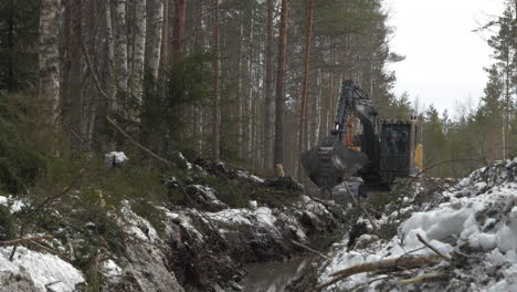 Bagger-Räumt-Verschneites-Waldgelände,-Schlammige-Spuren-Zwischen-Bäumen,-Mechanischer-Arm-In-Bewegung,-Bewölkter-Tag