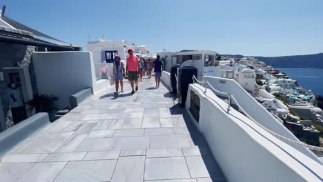 Couple's-On-Sidewalk-Near-Cliff-|-Oia-Santorini-Greece-Greek-Island-in-Aegean-Sea,-Travel-Tourist-Vacation-Immersive-Moving-Walk-Along-Crowds-Shopping-in-White-Marble-Cliffside-and-City,-Europe,-4K