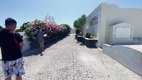 Turistas-Posando-Cerca-De-Flores-|-Oia-Santorini-Grecia-Isla-Griega-En-El-Mar-Egeo,-Viajes-Turísticos-Vacaciones-Inmersivas-Caminatas-En-Movimiento-Junto-A-Multitudes-De-Compras-En-Acantilados-De-Mármol-Blanco-Y-Ciudad,-Europa,-4k