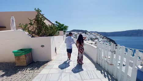 Women-Sitting-Looking-Over-Cliff-|-Oia-Santorini-Greece-Greek-Island-in-Aegean-Sea,-Travel-Tourist-Vacation-Immersive-Moving-Walk-Along-Crowds-Shopping-in-White-Marble-Cliffside-and-City,-Europe,-4K