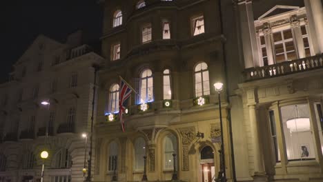 Cinematic-shot-of-architecture-of-a-vintage-building-with-flag-of-united-Kingdom-beside-a-busy-road-during-night-in-London,-England