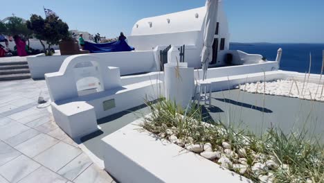 Distant-Woman-In-Blue-Dress-Posing-|-Oia-Santorini-Greece-Greek-Island-in-Aegean-Sea,-Travel-Tourist-Vacation-Immersive-Moving-Walk-Along-Crowds-Shopping-in-White-Marble-Cliffside-and-City,-Europe,-4K