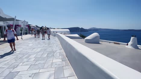 Menschen-Auf-Weißem-Bürgersteig-In-Der-Nähe-Von-Café-|-Oia,-Santorini,-Griechenland,-Griechische-Insel-Im-Ägäischen-Meer,-Reise,-Touristenurlaub,-Immersiver-Wanderspaziergang-Entlang-Der-Menschenmassen,-Die-In-Klippen-Aus-Weißem-Marmor-Einkaufen,-Und-Stadt,-Europa,-4k