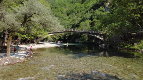 Touristen-Bestaunen-Die-Atemberaubende-Landschaft-Am-Flussufer-Des-Voidomatis,-Wobei-Die-Berühmte-Aristi-Brücke-Die-Schönheit-Der-Landschaft-Noch-Verstärkt