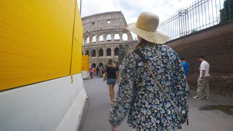 Rom-Immersiver-POV:-Umzug-In-Belebten-Straßen-Zur-Chiesa-Santi-Luca-E-Martina,-Italien,-Europa,-Gehen,-Wackelig,-4k-|-Frau-Mit-Hut-Geht-Mit-Einem-Schild-Vor-Dem-Kolosseum-In-Der-Nähe-Einer-Langen-Touristenschlange-Entlang