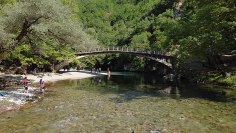 Touristen-Genießen-Die-Landschaft-Am-Ufer-Des-Flusses-Voidomatis,-Wo-Die-Berühmte-Aristi-Brücke-Zur-Schönheit-Der-Landschaft-Beiträgt