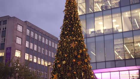 Top-down-cinematic-close-up-shot-of-a-huge-decorated-Christmas-tree-surrounded-by-wooden-fence-on-a-street-during-evening-in-Manchester,-England