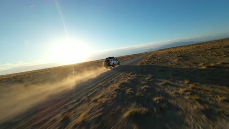 Low-cinematic-aerial-following-SUV-driving-into-the-sunset-on-scenic-desert-dirt-road,-Utah