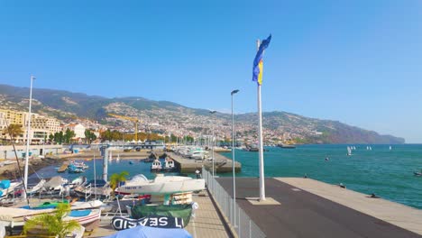 Ondeando-La-Bandera-De-Funchal-En-Portugal-Durante-Un-Día-Soleado-En-El-Puerto-Con-La-Playa-Y-La-Ciudad-En-La-Ladera-De-La-Montaña---Plano-Amplio