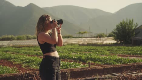 Woman-in-black-tank-top-raises-and-holds-camera-to-eye-in-front-of-farmland-on-Mediterranean-island