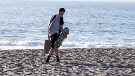 Un-Patinador-Llevando-Su-Tabla,-Una-Toalla-Y-Una-Bolsa-De-Papel-Por-La-Playa-En-Un-Día-Soleado