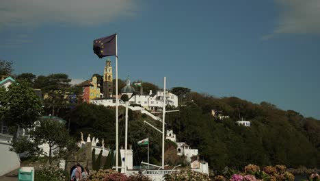 Bandera-De-La-Unión-Europea-Ondeando-En-Primer-Plano,-Dos-Banderas-Galesas-Con-La-Aldea-De-Portmeirion-Al-Fondo,-Gente-Caminando