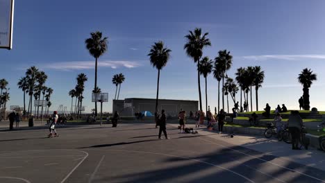 Gente-Pasando-El-Rato-Y-Montando-Patinetas-En-Las-Canchas-De-Baloncesto-Del-Paseo-Marítimo-De-Venice-Beach-Por-La-Noche-En-Los-Ángeles,-California,-Estados-Unidos---Plano-Amplio