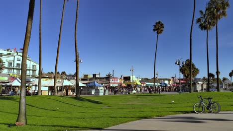 Paseo-Marítimo-De-Venice-Beach,-Gente-Caminando-Por-La-Franja-Al-Fondo-Con-Tiendas-Y-Puestos-En-Una-Tarde-Soleada-En-Los-Ángeles,-California,-EE.UU.---Toma-Estática-Portátil