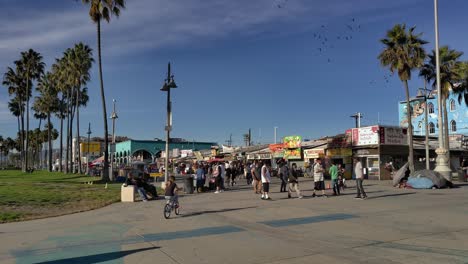 Gente-Caminando-En-El-Paseo-Marítimo-De-Venice-Beach,-Tiendas-Y-Puestos-Y-Palmeras-Y-Parques-Cubiertos-De-Hierba-En-El-Fondo-En-Los-Ángeles,-California,-EE.UU.