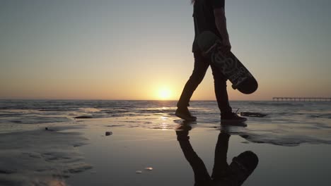 Toma-Cinematográfica-En-Cámara-Lenta-De-Un-Joven-Con-Patineta-Caminando-Sobre-Acantilados-Cerca-Del-Océano-Durante-La-Puesta-De-Sol.