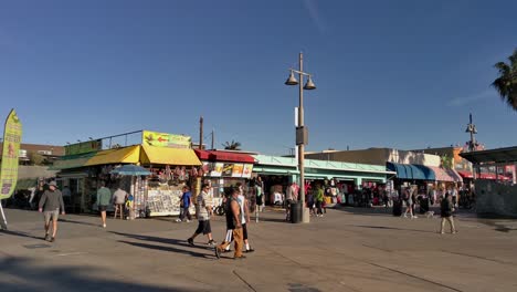 People-Leisurely-Walking,-Biking,-Rollerblading,-And-Skateboarding-At-Ocean-Front-Walk-Next-To-Gift-Shops-And-Food-Stalls-Near-Venice-Beach-In-Los-Angeles,-California,-USA