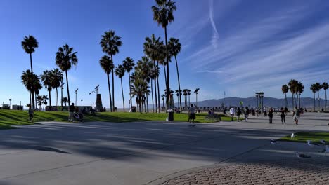 People-passing-by-on-skateboards-on-crowded-Venice-Beach-Boardwalk-during-Covid-19-with-skatepark-and-art-sculptures-in-the-background-in-Los-Angeles,-Califonia,-USA