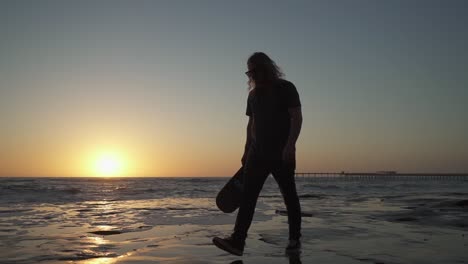 Cinematic-slow-motion-shot-of-young-male-holding-skateboard-on-cliffs-near-the-ocean-during-sunset