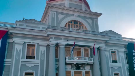 Un-Hombre-Camina-Frente-A-Un-Edificio-Gubernamental-Que-Tiene-Una-Gran-Bandera-De-Cuba-Y-Otras-Banderas-En-La-Plaza-Principal-De-La-Ciudad-De-Cienfuegos,-Cuba.