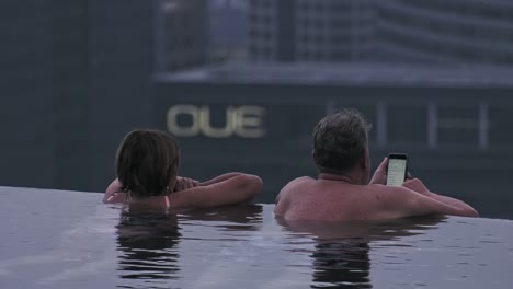 Pareja-De-Mediana-Edad-Mirando-Hacia-Atrás-Descansando-En-El-Borde-De-La-Piscina-Infinita-En-Marina-Bay-Sands,-Singapur---Toma-Estática-Media-Larga