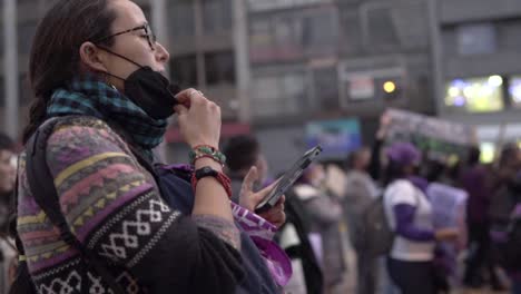 A-woman-is-wearing-a-black-mask-and-watching-her-cellphone-during-a-protest-in-the-International-Women's-day-in-Quito,-Ecuador,-People-on-the-back-singing-and-holding-up-sigs