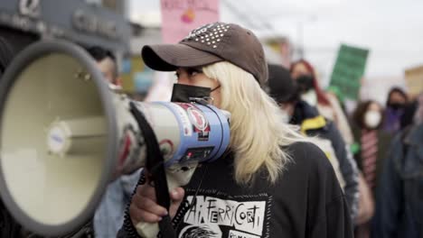 A-woman-wearing-a-mask-and-a-hat-is-holding-up-a-megaphone-and-speaking-to-motivate-the-women-marching-with-her-during-the-protest-in-the-International-Women's-day-in-Quito,-Ecuador