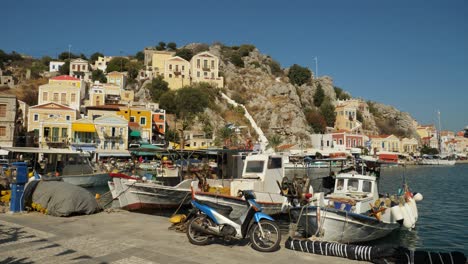 Spiel-Des-Sonnenlichts-Auf-Den-Booten,-Die-Im-Hafen-Der-Insel-Symi-Festmachen,-Malerischer-Hintergrund-Der-Stadt-Auf-Dem-Hügel