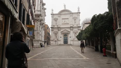 Disparo-De-Inclinación-Lenta-En-La-Plaza-Junto-A-La-Iglesia-De-San-Roque,-Hombre-Caminando-Con-Una-Máscara-Protectora-Negra,-Dos-Días-Después-Del-Carnaval-Cancelado